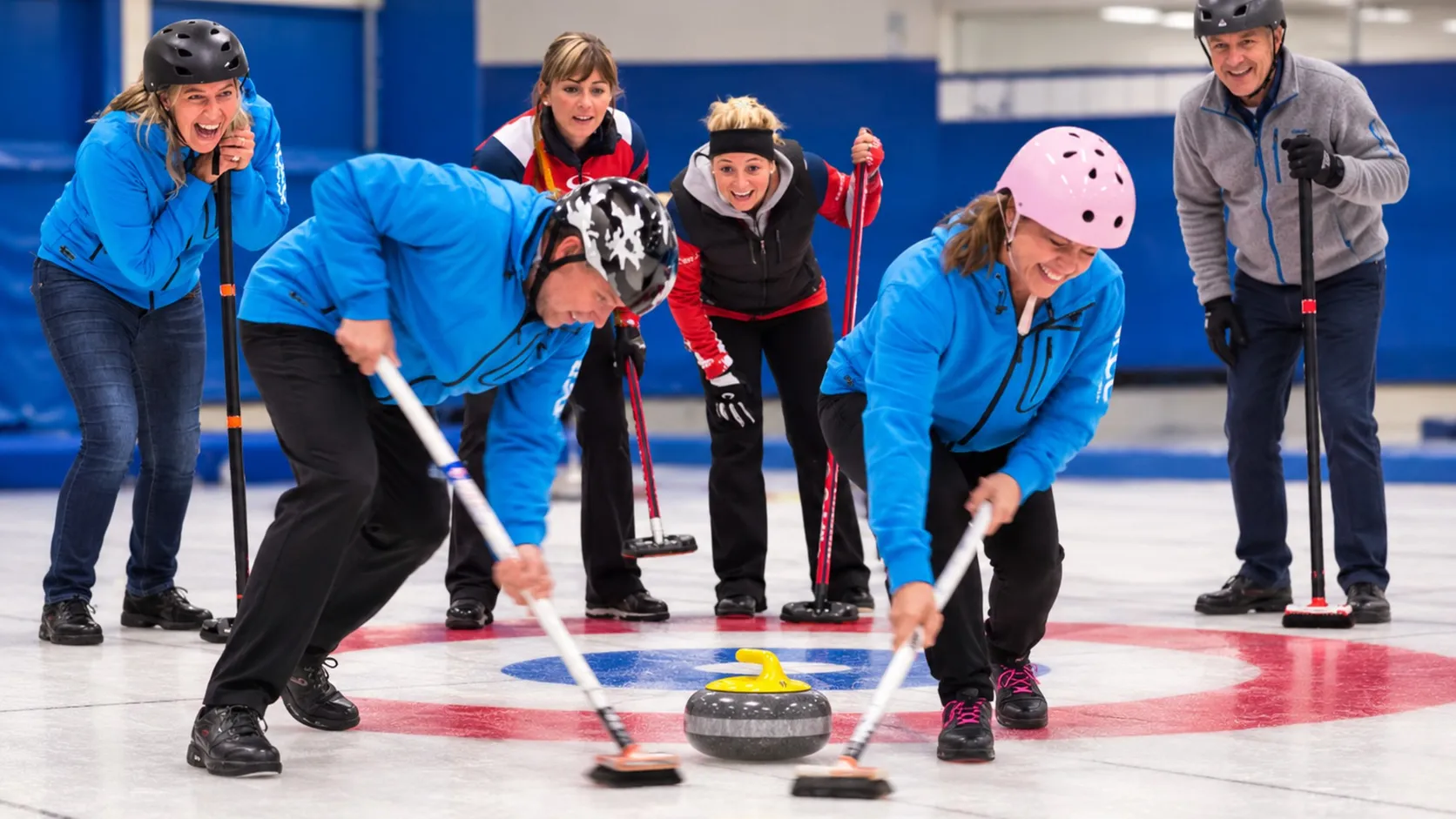 Ett glatt lag spelar curling inomhus, två deltagare sopar isen framför stenen medan resten av laget hejar.